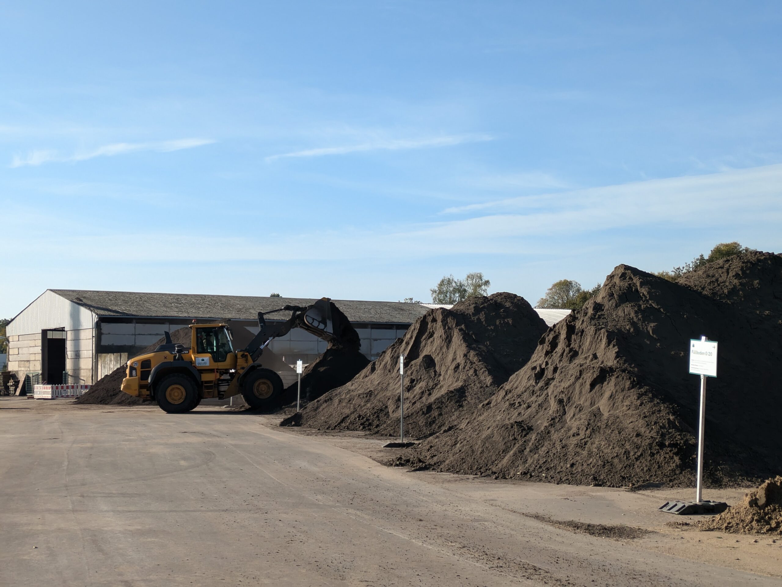 Gelber Radlader hebt Erde von einem großen Erdhaufen auf einem Baustellenareal. Im Hintergrund ist ein Lagergebäude sichtbar.