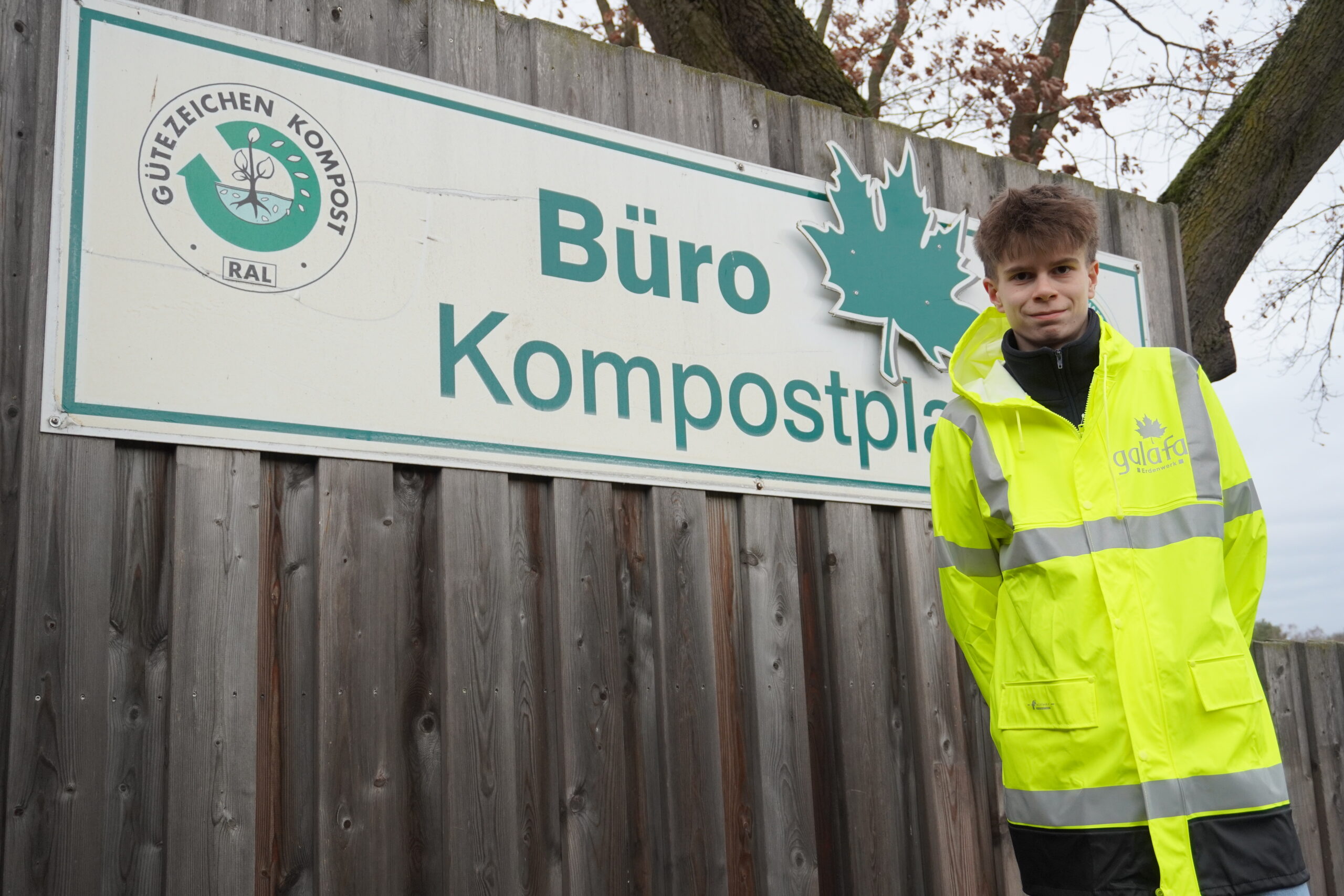 DSC00059 Junger Mann in leuchtend gelber Arbeitsjacke mit reflektierenden Streifen steht vor einem Schild mit der Aufschrift "Büro Kompostplatz" und einem Gütezeichen für Kompost.