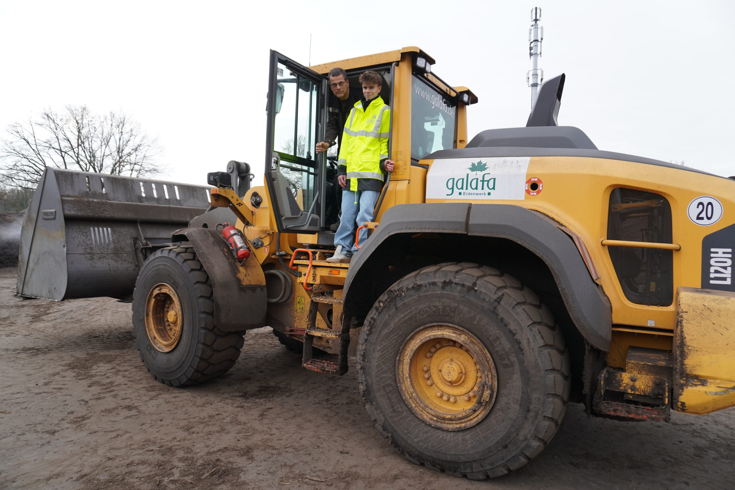 DSC00064 Zwei Personen stehen auf dem Fahrersitz eines gelben Radladers der Marke Galafa. Eine Person trägt eine leuchtend gelbe Sicherheitsjacke, während die andere in normaler Kleidung gekleidet ist. Im Hintergrund ist die Schaufel des Radladers sowie eine baumlose Landschaft sichtbar.