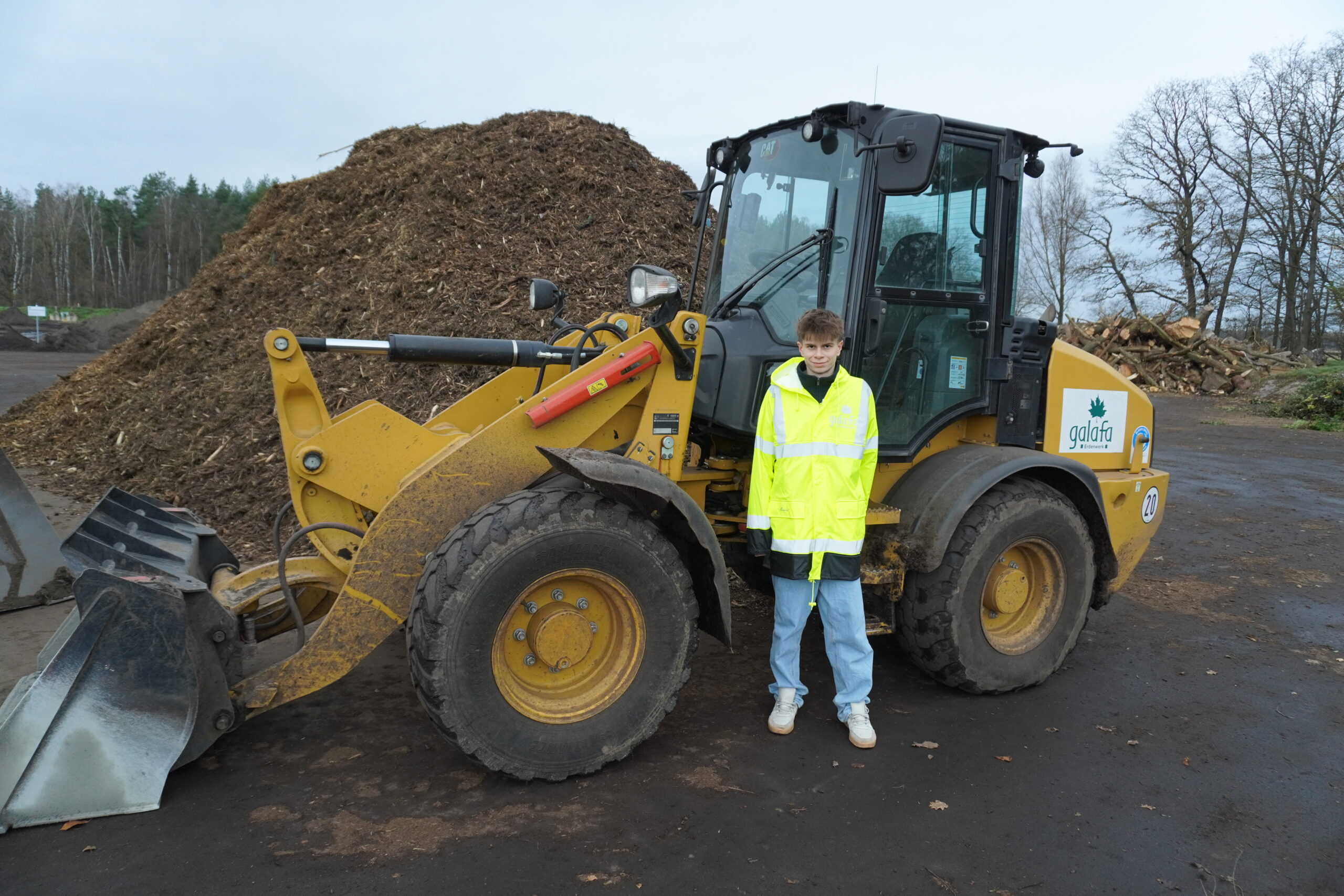 DSC00069 Junge in gelber Sicherheitsjacke steht neben einem gelben Radlader auf einem Recyclinggelände. Im Hintergrund ist ein Haufen organischer Abfälle sichtbar.