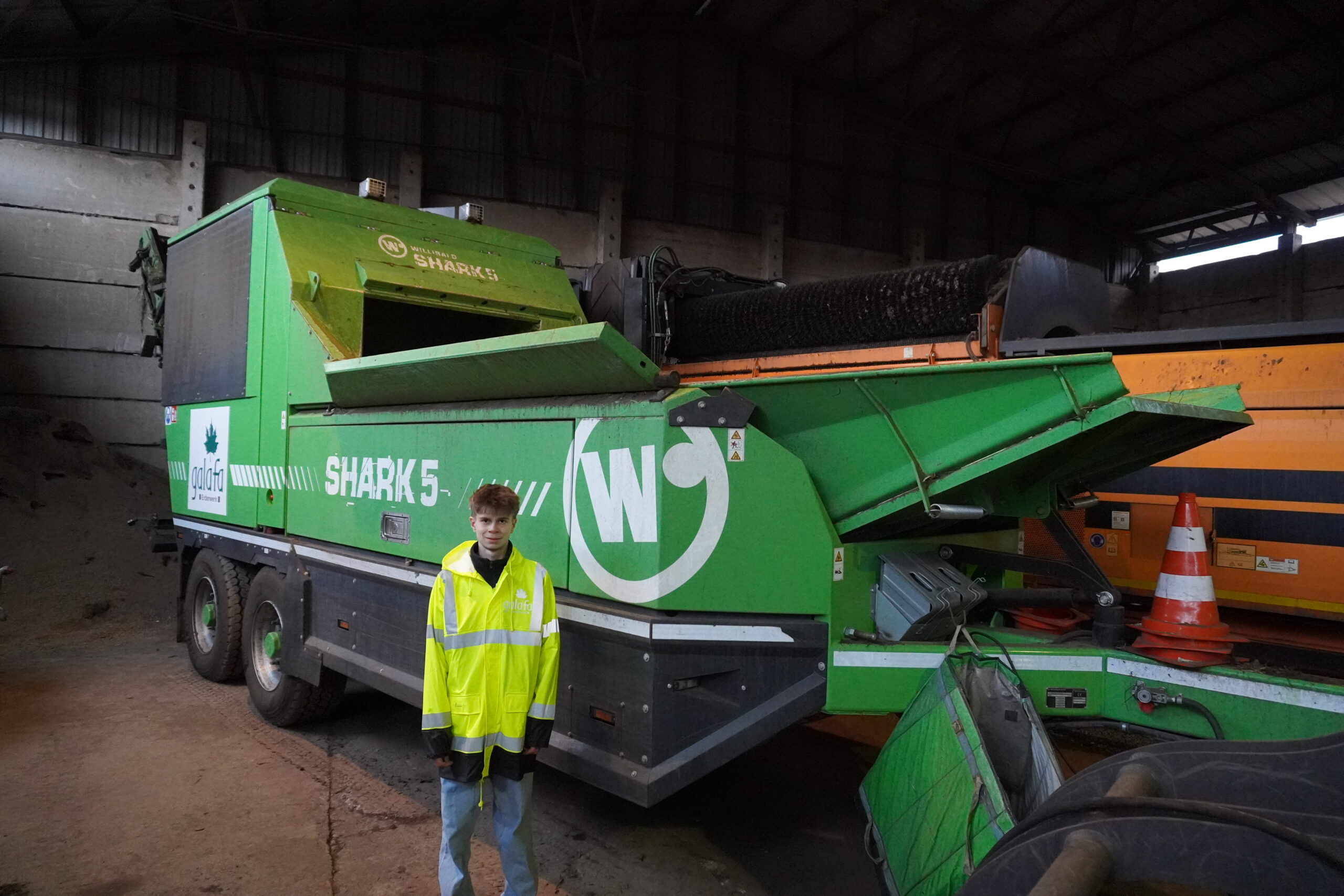DSC00079 Grüner Schredder "Shark 5" neben einer Person in reflektierender Jacke, im Hintergrund eine Lagerhalle.